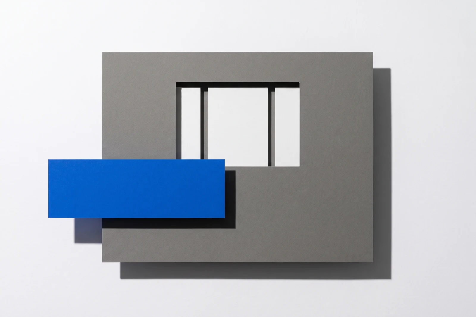 Une fenêtre rectangulaire découpée dans une plaque de carton gris laisse voir des cartes dessous, ombres dures projetées vers le bas à droite.