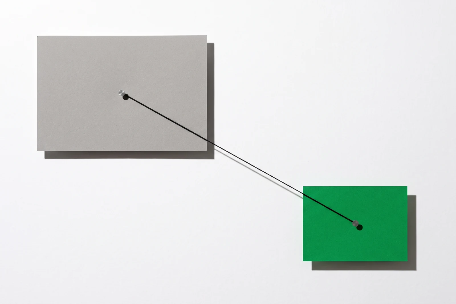 Une plaque de carton gris punaisée en haut à gauche et une carte de papier vert punaisée en bas à droite, reliées par un seul fil de coton noir tendu en diagonale, ombres dures projetées vers le bas à droite.