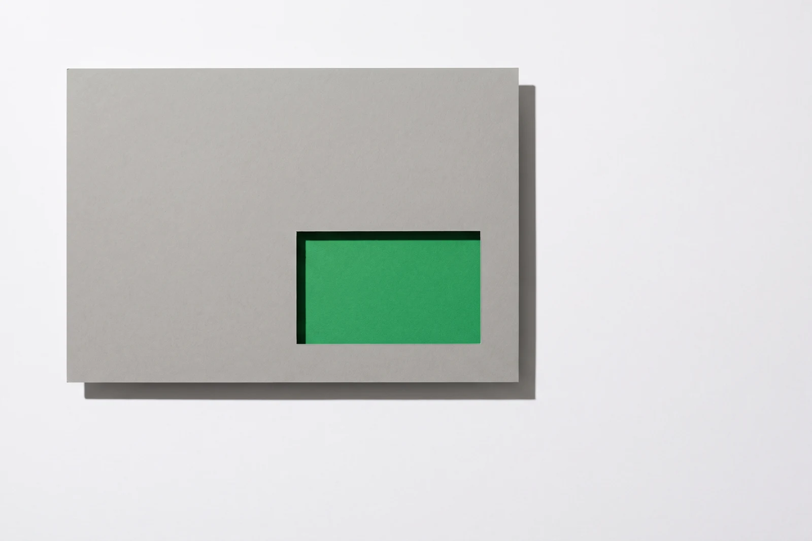 Une plaque de carton gris posée à plat, percée d’une fenêtre rectangulaire près de son coin inférieur droit qui laisse voir une feuille de papier vert dessous, ombres dures projetées vers le bas à droite.