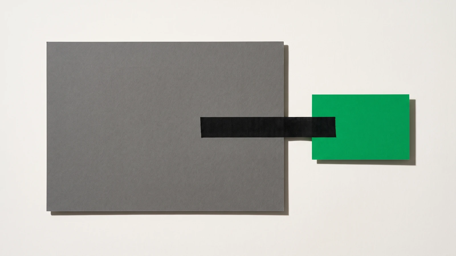 Une large plaque de carton gris et une petite carte de papier vert posées à plat, reliées par une seule bande de toile noire tendue en travers du vide qui les sépare, ombres dures projetées vers le bas à droite.